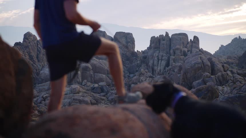 A man and his dog stand on rocky ground overlooking a desert canyon at sunset