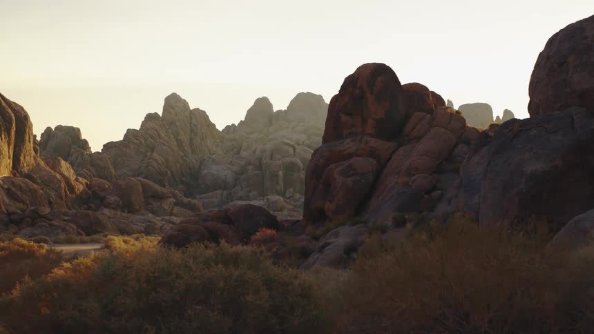 A small figure of a man wearing dark clothes climbs over a large reddish brown rock formation in a vast dry desert landscape with rugged boulders and scrub brush at sunset