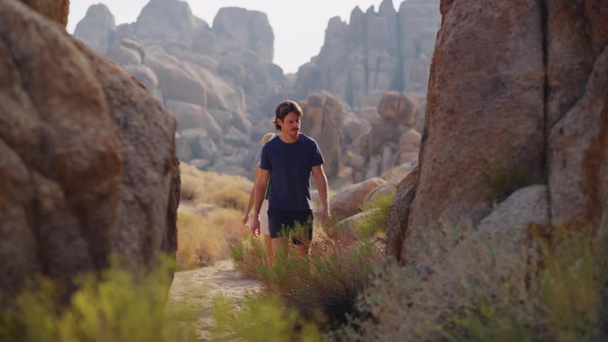 A couple walks along a sunny narrow hiking trail flanked by arid brush and tall jagged rock formations in the distance the pair dressed in shorts and tshirts walk with a focused steady pace as they navigate the rocky desertlike terrain