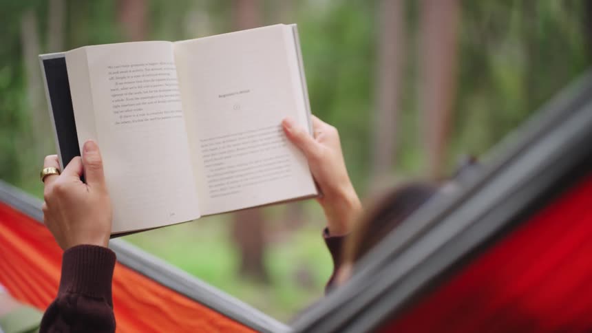 A woman lying in a hammock outdoors reading a book