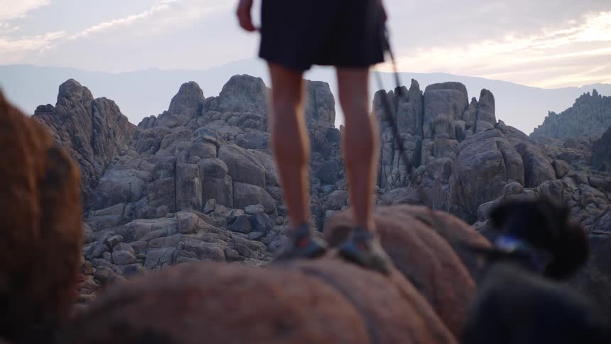 A man hikes on a rocky formation at midday standing on a cliff while holding a dog on a leash