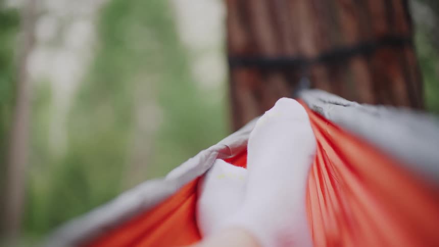 A close view from inside an orange hammock shows a persons feet wearing white socks resting in the fabric with a blurry green forest background and tree trunk visible