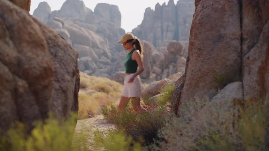 A man and a woman walk away from the camera passing between two large foreground boulders along a path with a field of scrub brush and imposing rugged rock formations in the background