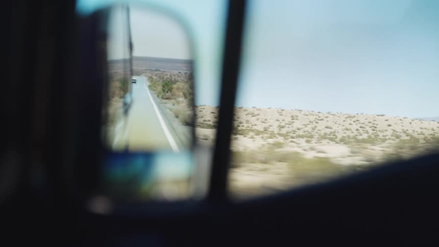 A car travels down a highway on a sunny day road trip with the side mirror reflecting the empty desertlined road behind
