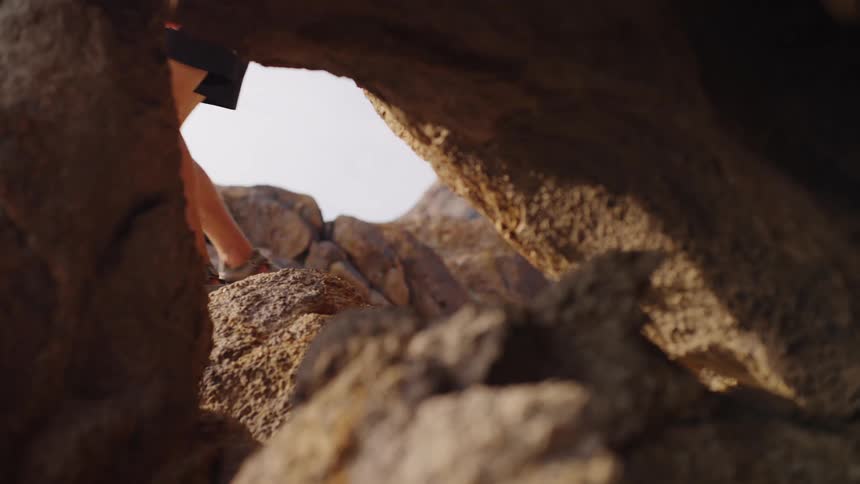 The video is shot through a natural rock arch or opening framed by rugged brown boulders in the foregroundin the middle ground a persons lower body wearing dark shorts and shoes walks across the sunlit rocky terrain and then moves out of