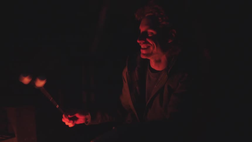 A man in front of a campfire holding a skewer with marshmallows he puts the marshmallow in the fire and then blows on it