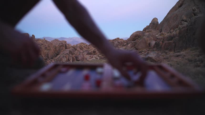 Two people sit opposite each other and move game pieces across a blurry backgammon board in the foreground silhouetted against a vast rugged landscape of large boulders and distant mountains under a twilight sky