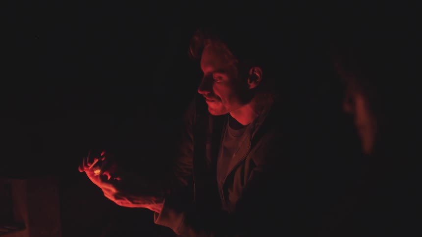 A man with short hair is dimly lit by a fire or small light source that casts an orange glow on his face and hands as he sits in the dark