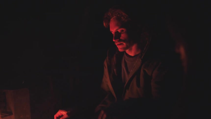 A man with short hair is dimly lit by a fire that casts an orange glow on his face as he sits in the dark and looks downward