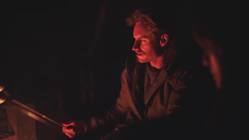 Two men sit by a campfire lit by flickering flames at night