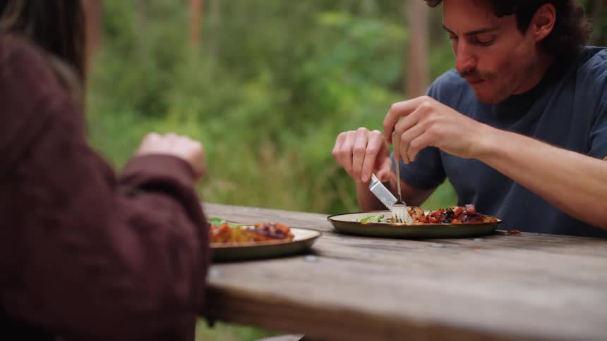 Two people eat together at a wooden table in a forest setting