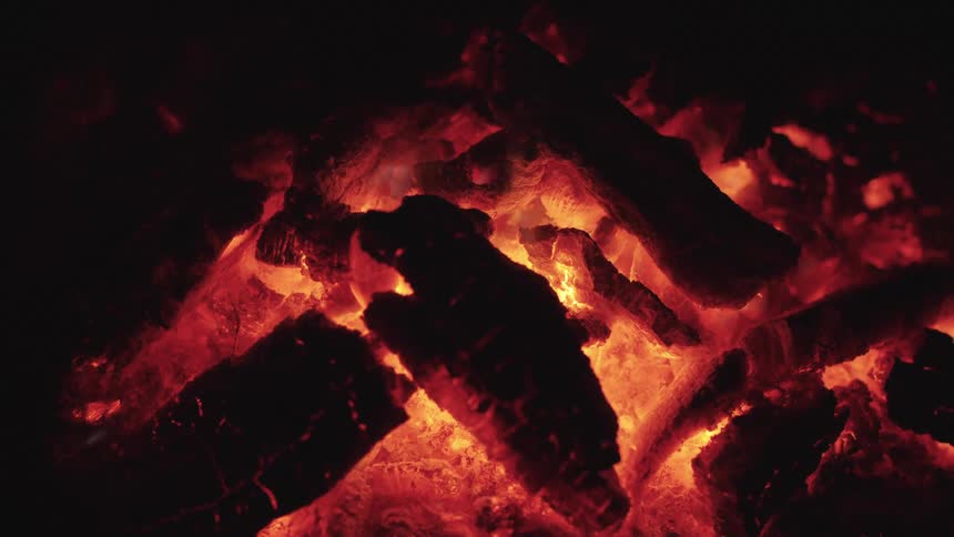 A closeup of several dark wood or charcoal pieces shows them glowing with an intense steady red and orange heat surrounded by darkness