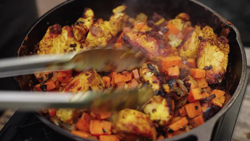 A pair of metal tongs stirs and flips pieces of seasoned chicken and chopped vegetables that are sizzling and steaming in a black cast iron pan on a stovetop