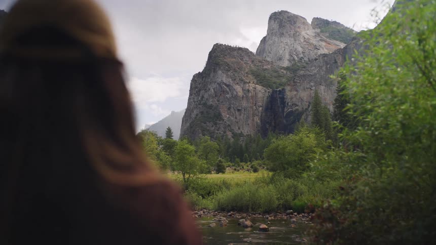 The video shows a person wearing a tan baseball cap and a dark shirt from behind looking out over a stunning natural landscapein the distance towering granite cliffs or mountainous rock formations rise sharply above a lush green valley with a stream