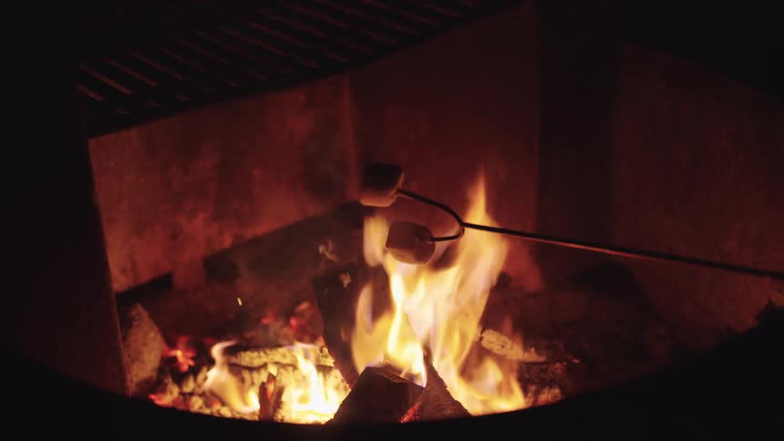 Two marshmallows on a roasting stick are held over a crackling campfire in a fire pit