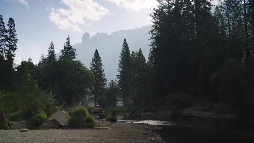 A man and woman are walking their dog on a leash through a forest in the mountains along a river