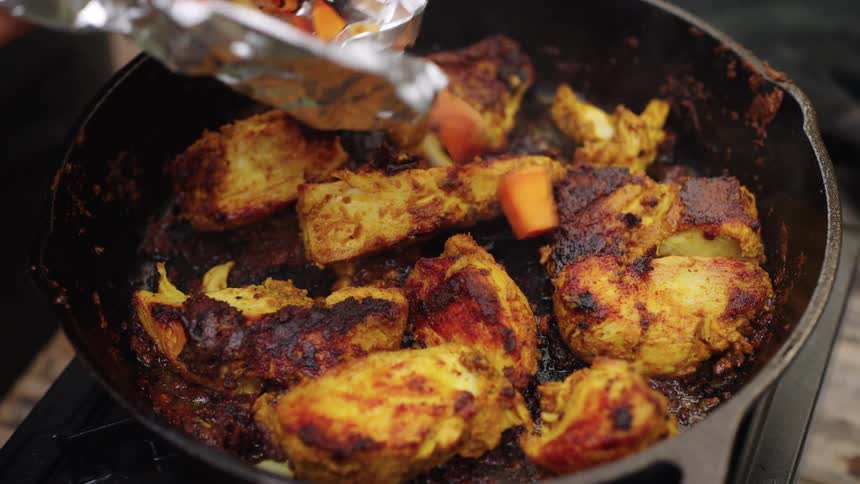 A person fries pieces of chicken in a pan and adds sliced carrots and onions