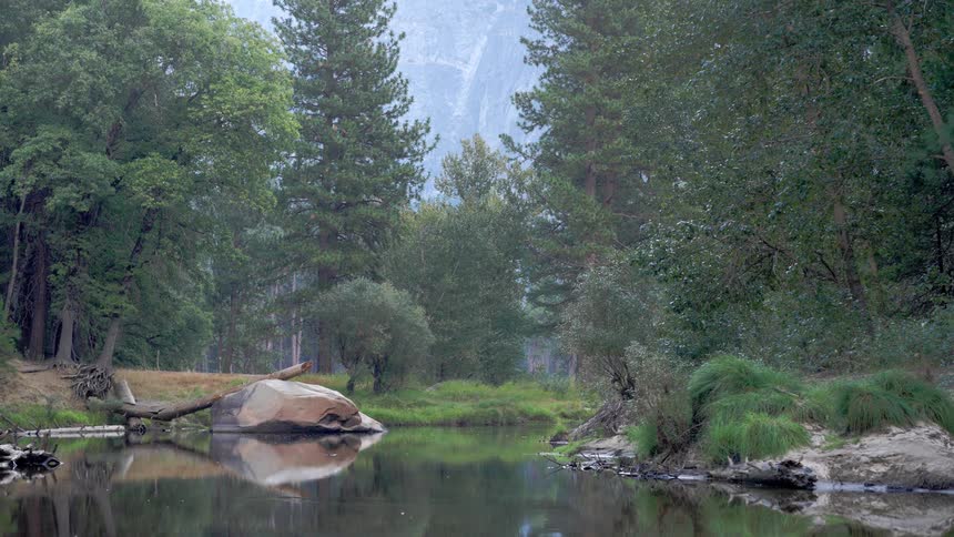 A slowmoving body of water reflects the surrounding landscape a large lightcolored rock sits near the center and tall evergreen trees line the banks leading up to sheer cliffs shrouded in haze in the distance