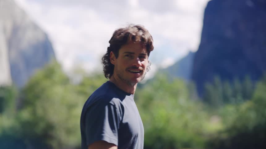 Man smiles outdoors with mountains in the background on a sunny day