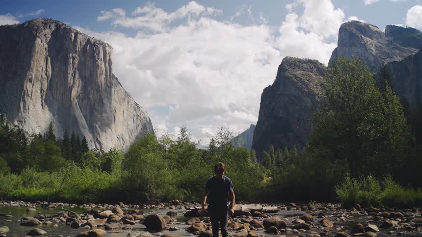 A young man in dark clothes stands in a rocky riverbed stretches his arms high and looks up at the towering granite cliffs and green forests of yosemite valley on a bright cloudy day