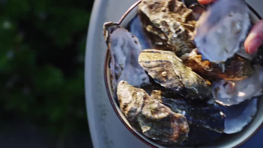 A bowl is filled with shucked and halfshelled oysters and a persons hand put another one on top and a blurred background fills the scene