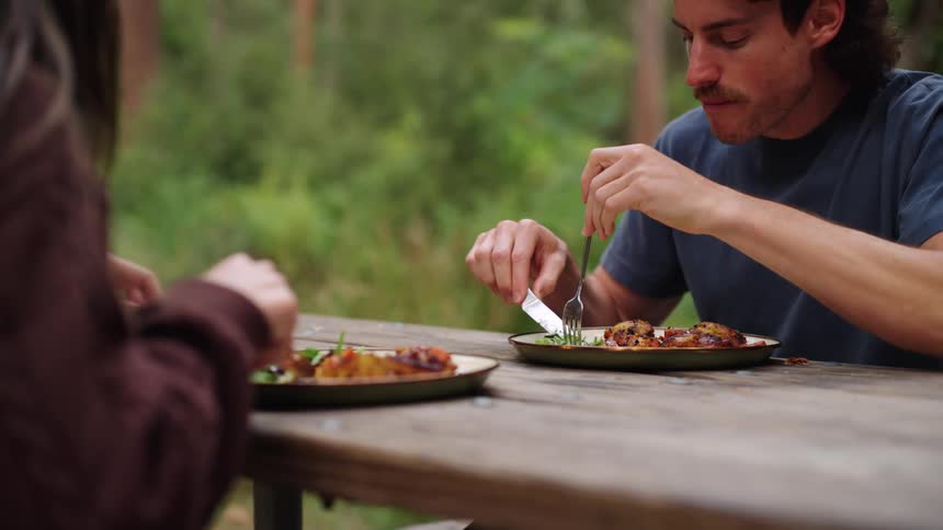 A man sitting at a table outdoors eating with a fork and knife with a woman in front of him also eating