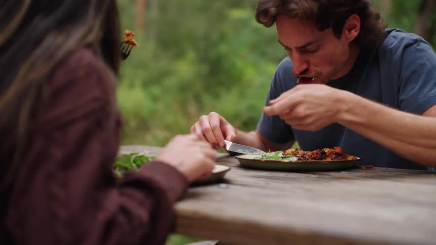 A man sitting and eating from a plate with a fork and knife with a woman in front of him also eating