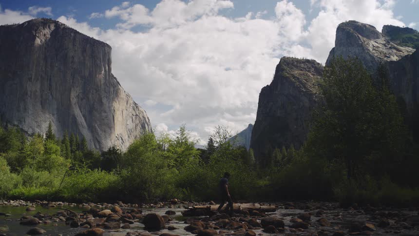 A man crosses a shallow rocky stream beneath tall mountains and greenery
