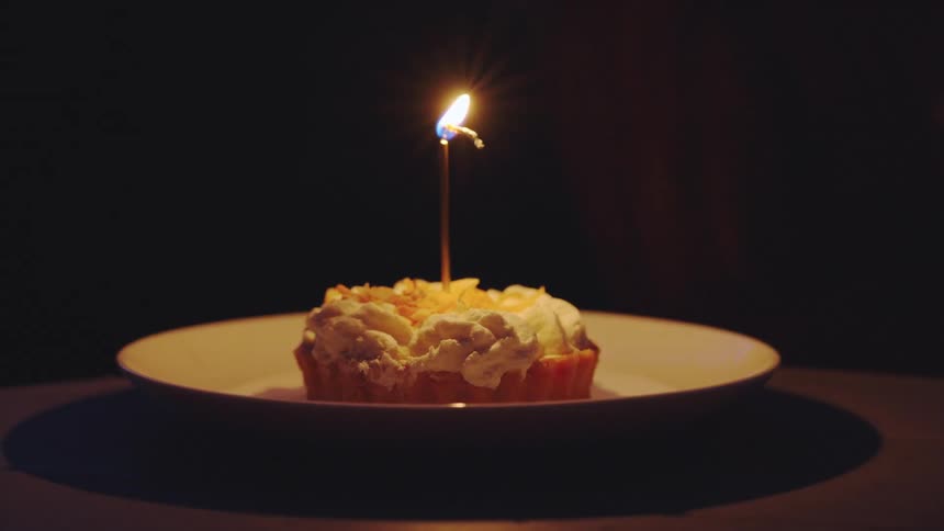 A personal desert pie is placed on the table its covered in whipped cream and has a lit candle on it someone off screen blows out the candle