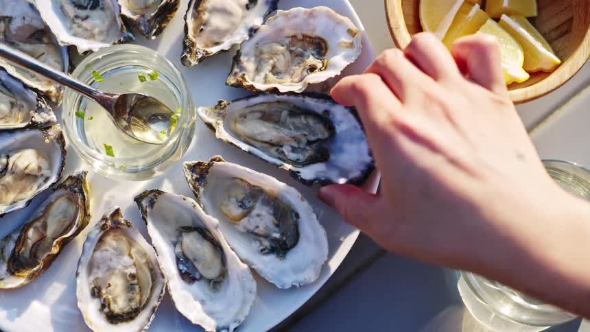 A hand selects a fresh oyster from a platter served with lemon wedges and a vinaigrette