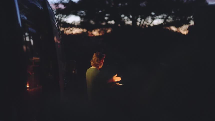 A young man steps out of a dark vehicle into a dark outdoor setting at dusk holding a small burning candle on a plate then walks away from the camera