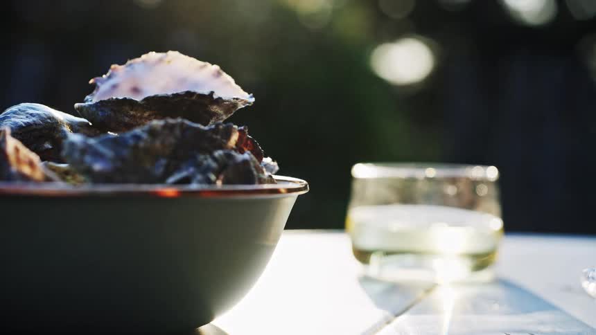 A dark green bowl filled with dark shellfish rests on a bright surface a hand enters the frame and lifts one shell and a glass of liquid sits out of focus in the background all bathed in bright sunlight
