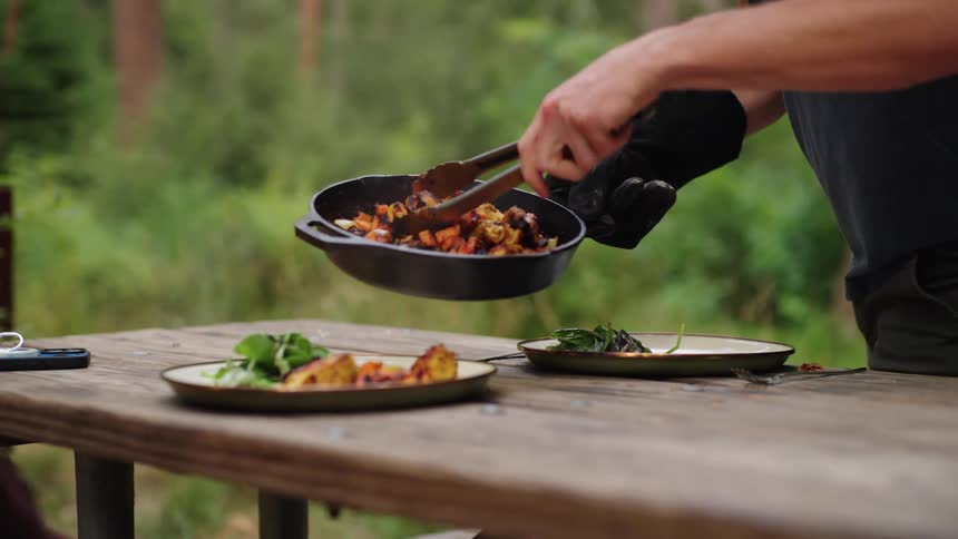 Person serving food from a skillet onto plates on a wooden outdoor table