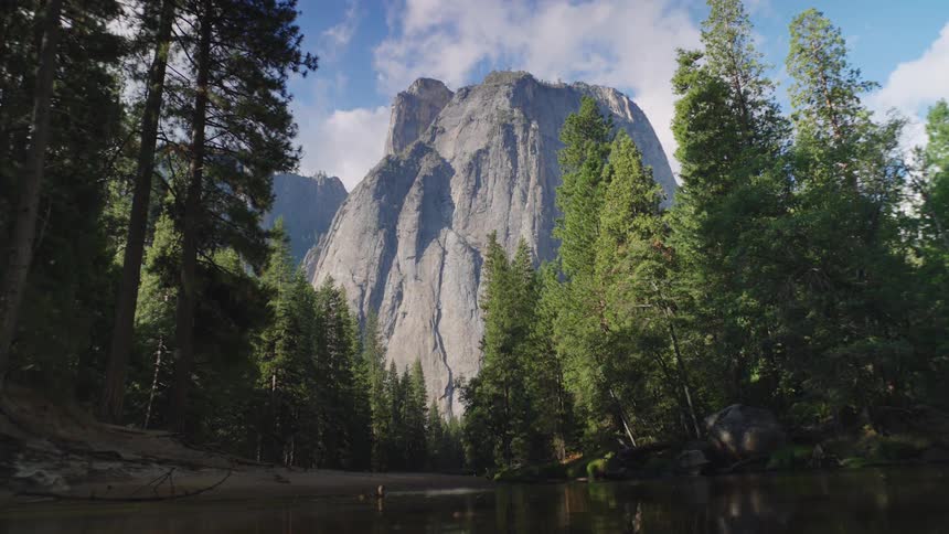 Towering mountain surrounded by pine trees reflects on calm water under a partly cloudy sky