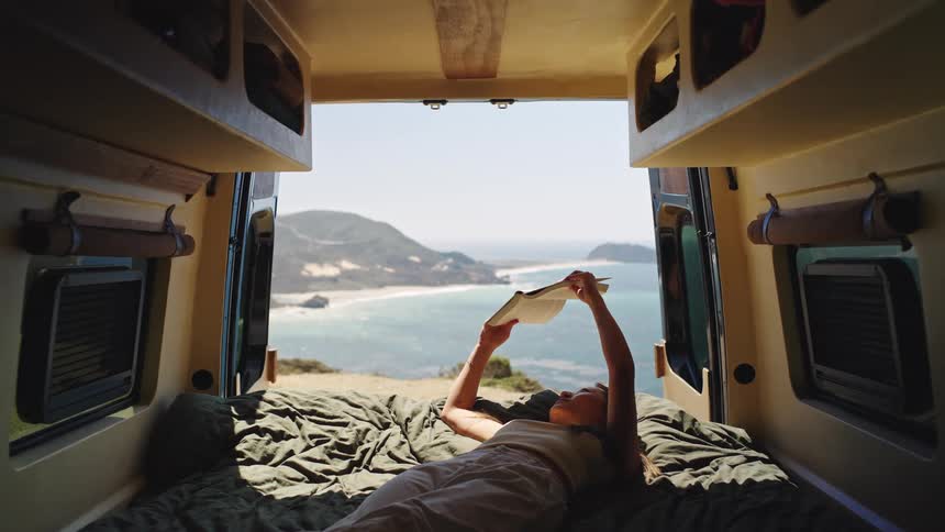 A woman is laying in the bed in a campervan and reading a book