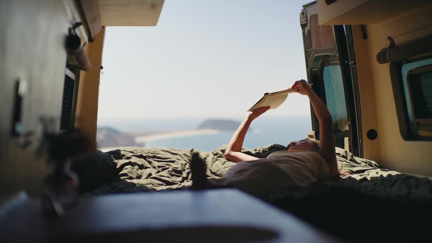 A woman lies on their back on a bed inside a vehicle holds a book open over their head while reading and through the vehicles rear opening a body of water and distant land are visible under bright sunlight