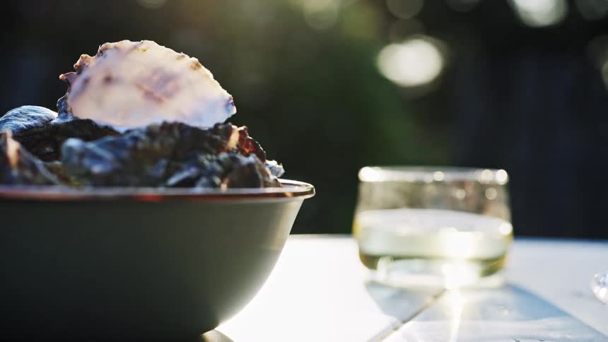 A close view shows a dark bowl filled with mussels and an empty shell sitting next to a short glass of white wine on a sunny outdoor table