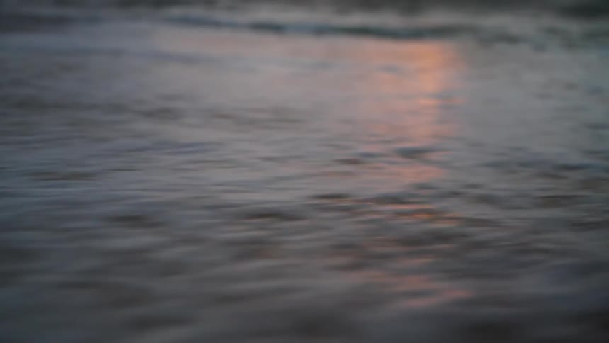 The video is a lowangle closeup of a foamy wave gently rolling onto a sandy beach the shot has a dark muted color palette with the water and sand tones blending into the low light conditions