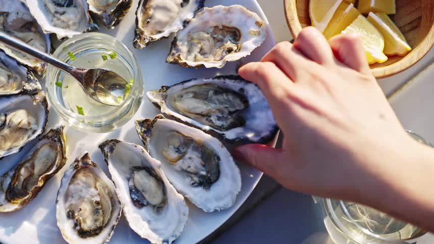 A table with a plate filled with oysters as a person picks up a piece to enjoy at midday