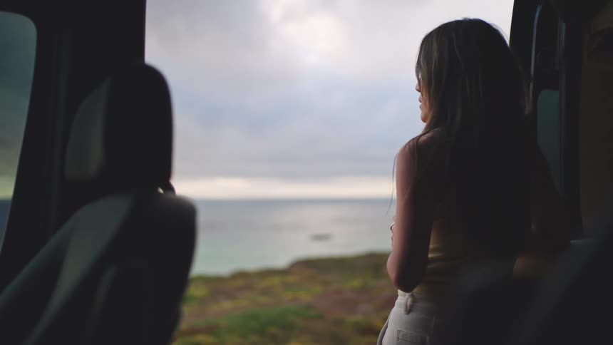 The video shows a woman seen from the back and inside a vehicle standing by an open door or window looking out at the ocean vistashe is holding a white and yellow travel mug or container and takes a moment to drink from it while enjoying the