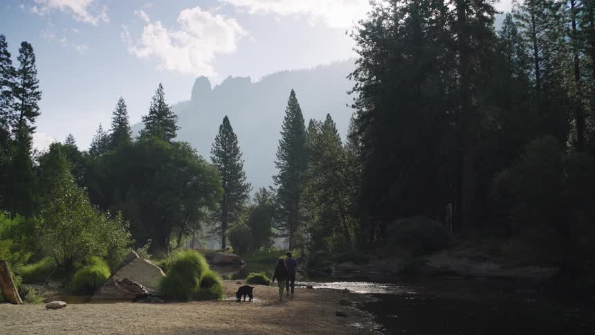 Couple walks with a dog near a river surrounded by forest and mountains
