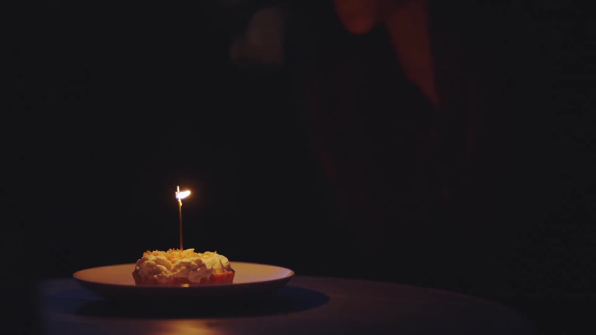A person places a cake with a single candle on a plate in front of a woman in a dark room and as she blows out the candle the room is plunged into complete darkness