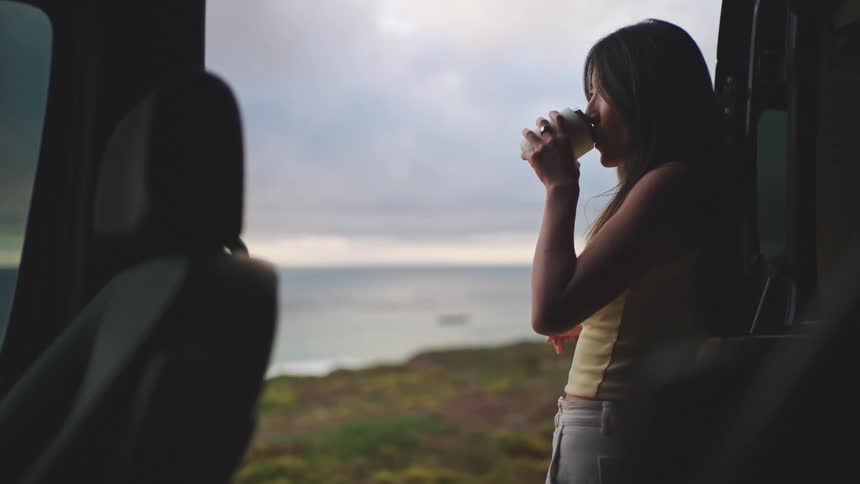 Woman standing at open side door of vehicle parked in front of landscape with water cloudy sky and low vegetation