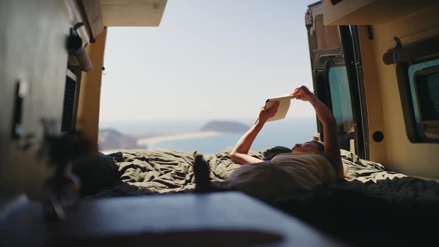 A woman lies on their back on a darkcovered mattress inside a vehicle holding a book with both hands above the face and the open rear of the vehicle frames a bright background view of a beach