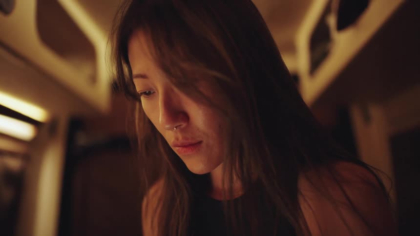 The video is a medium closeup of a young woman with long dark hair that partially obscures her face and she has a nose piercingshe is looking downwards intently possibly at a device or object out of frame in a setting with warm dim ambient light