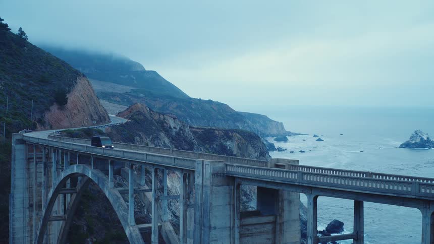 A coastal bridge stretches over the ocean beside rugged cliffs under a cloudy sky