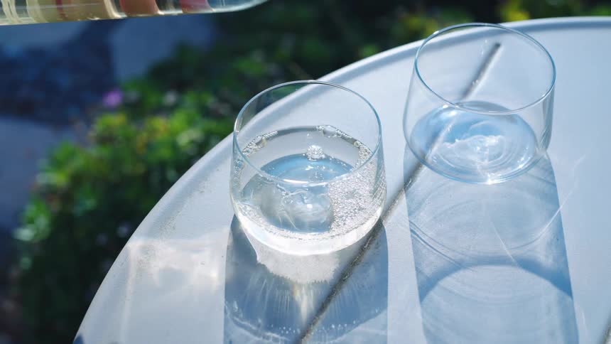 Hand of a white person pouring liquid from a glass bottle into two glass cups over a white table outdoors during day time
