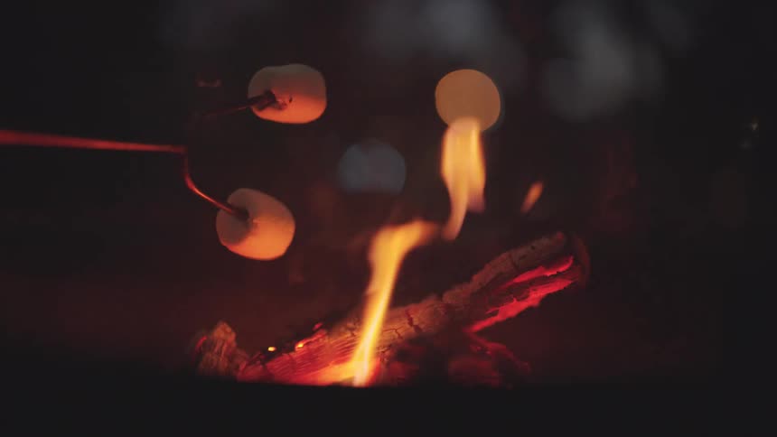 A closeup of two marshmallows roasting over a campfire at night