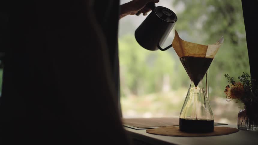 A person holding a kettle and pouring hot water into a glass jar with a coffee filter while in the background a woman wearing a cap is reading a magazine