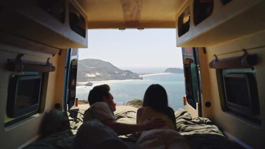 A man and a woman lie in the back of a van on a bed facing away from the camera they look out the wideopen rear doors and the doors frame a large view of a bright ocean coastline and rolling hills
